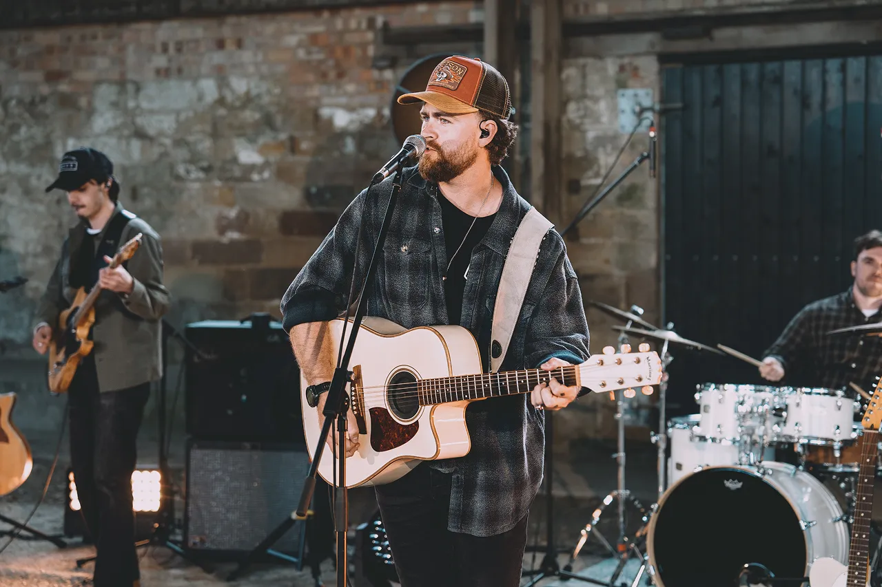Man in a cap playing acoustic guitar and singing into a microphone with band members in the background.