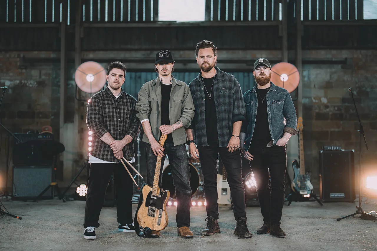 Four men in casual clothing standing in a music studio with guitars and drums in the background.